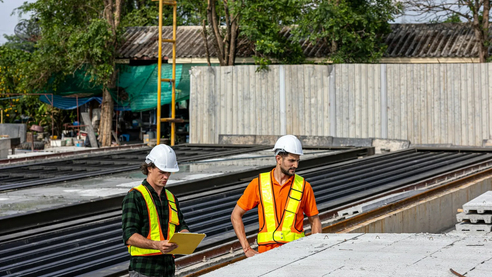Two workers in hard hats and high-visibility vests checking a clipboard on a construction site
